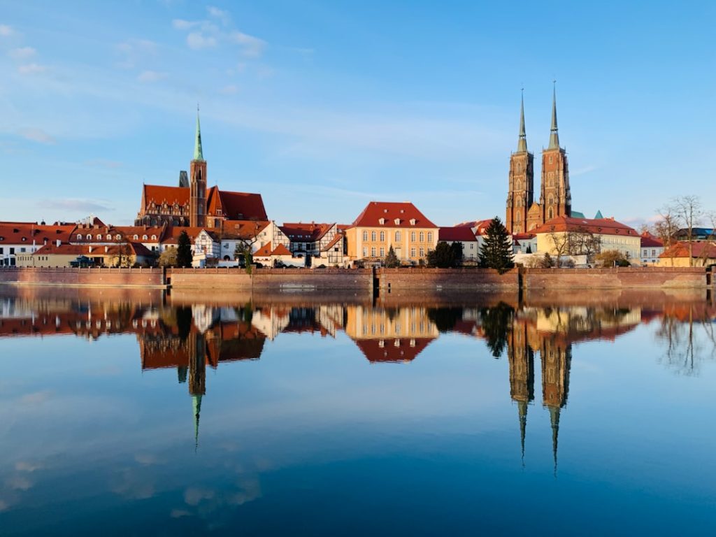 Architecture et berges à Wrocław Façades, toitures rouges et tours d’une église se reflétant dans une étendue d’eau calme sous un ciel bleu