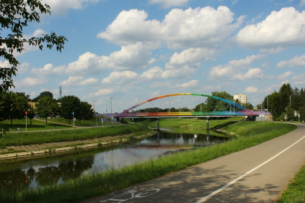 Pont aux arches colorées traversant un cours d’eau, avec une promenade, des pelouses et des arbres sur les berges