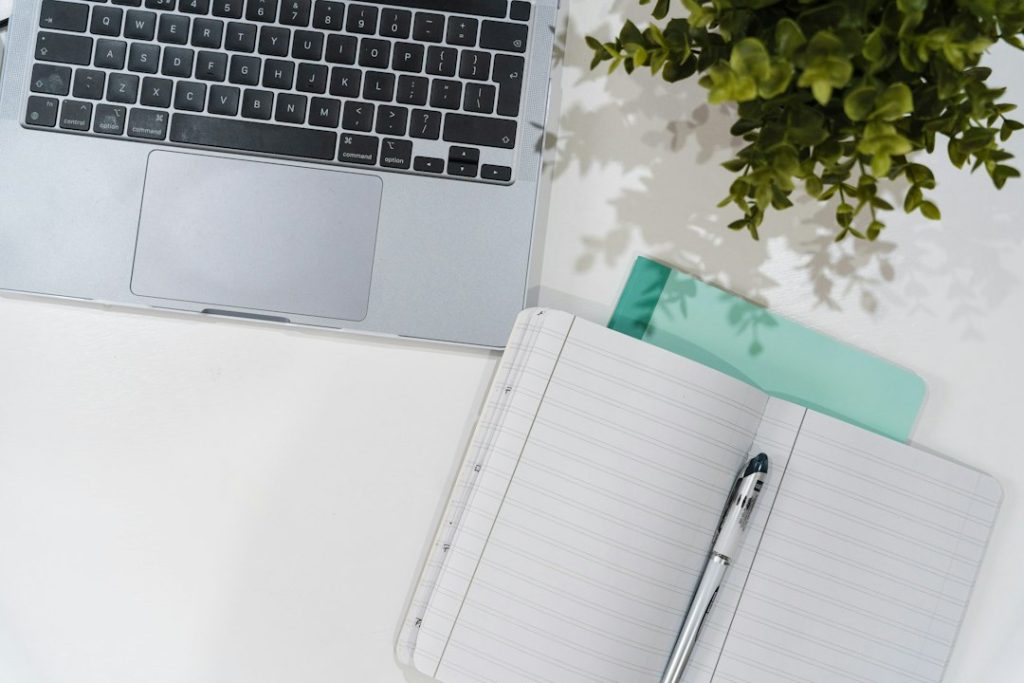 Carnet à pages quadrillées ouvert à côté d’un ordinateur portable, d’un stylo et d’une plante sur une table claire